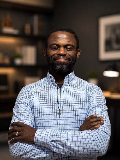 Riches Adejoro smiling with arms crossed, wearing a dark gray sweater and a cross pendant. A confident and inspiring leader in faith-based teachings and digital strategy